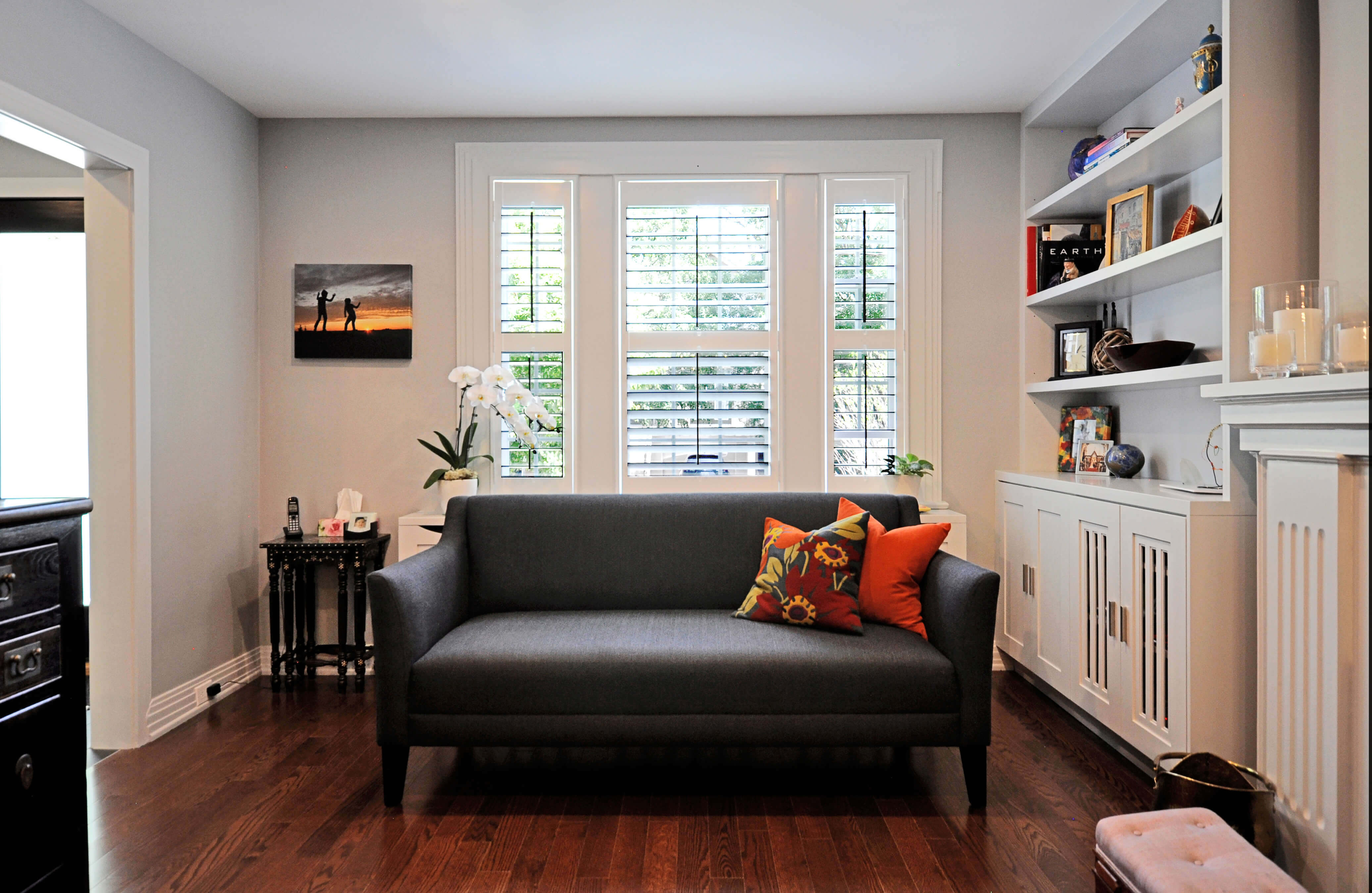 Living room. Grey couch with red accent pillows under a window. The window has white California shutters. Wood floors. Built in white floor to ceiling shelves.